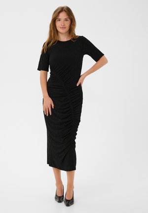 Femme debout, main sur la hanche, portant une robe midi noire froncée et des talons hauts noirs sur un fond blanc uni.