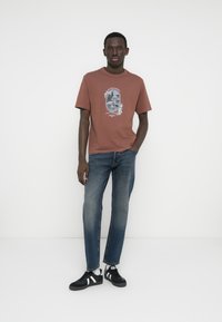 Κοντομάνικη μπλούζα σε σκουριά χρώμα με τυπωμένο σχέδιο, συνδυασμένη με μπλε τζιν παντελόνι κανονικής γραμμής και μαύρα αθλητικά παπούτσια με λευκές λεπτομέρειες.