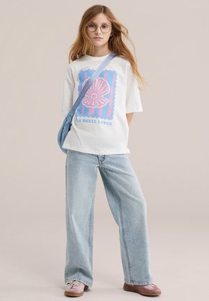 Jeune personne portant des lunettes, un t-shirt blanc avec un motif de coquillage, un jean large bleu clair, des chaussures roses et un sac bandoulière bleu.