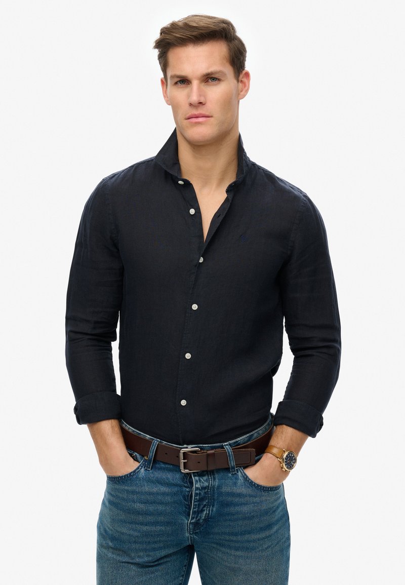 Navyblauwe linnen blouse met een button-down kraag, lange mouwen en een subtiel logo. Gecombineerd met een blauwe spijkerbroek en een bruine leren riem.