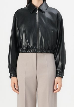 Kvinde iført cropped sort læderjakke med lynlås og lange ærmer og beige højtaljede bukser mod en ensfarvet baggrund.