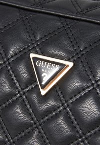 Borsa a tracolla nera trapuntata con una placca triangolare in oro e nero con il logo "GUESS U.S.A." e un design a forma di punto interrogativo. Materiale sintetico testurizzato.
