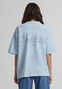 Camiseta de gran tamaño en azul claro con la palabra "Prohibido" en un estilo de escritura curvada en la parte posterior. Material de algodón con una textura suave y mangas cortas.