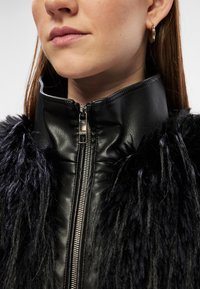 Casaco de pele preto com acabamento em pelo sintético, gola alta e detalhe em zíper prateado. Primeiro plano da área do pescoço e fecho em zíper.