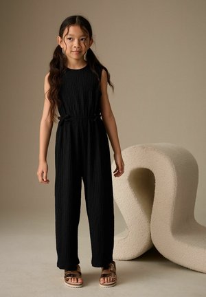 Junges Mädchen mit langem, welligem Haar, das einen schwarzen ärmellosen Overall und metallische Sandalen trägt und neben einer großen, cremefarbenen, strukturierten Skulptur steht.