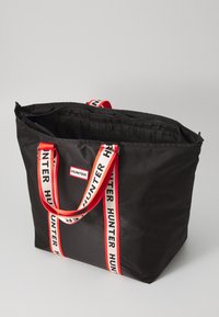 Bolsa de tote negra de nailon con asas de marca en rojo y blanco, cierre de cremallera y un interior espacioso. Presenta un logotipo llamativo en el panel frontal.