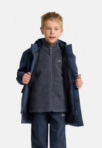 Berniukų striukė su tamsiai mėlynu vandeniui nelaidžiu išoriniu sluoksniu ir pilku vilnos vidiniu sluoksniu, su pilnu užtrauktuku ir kišenėmis, dėvima atidaryta.