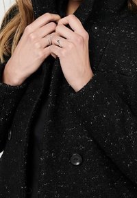 Musta tekstuuriga mantel koos kraega, millel on kaks suurt musta nuppu, käed on asetatud kaela juurde, näidates sõrmedel hõbedasi rõngaid.