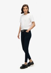 T-shirt blanc à manches courtes associé à un jean slim bleu foncé. Le modèle porte une ceinture noire et des mocassins noirs ornés d'un nœud.