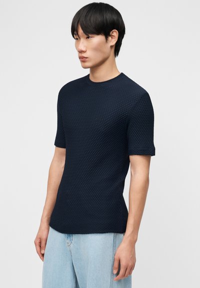 Νεαρός με μαύρα μαλλιά που φορά υφασμένο μπλε σκούρο κοντομάνικο πουκάμισο και ανοιχτόχρωμο μπλε τζιν, στέκεται μπροστά σε απλό φόντο.