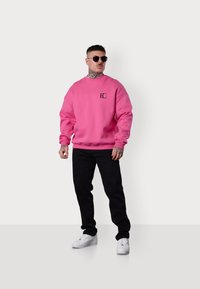 Un sweat-shirt rose avec un petit logo noir sur la poitrine, associé à un pantalon noir et des baskets blanches, porté par une personne aux cheveux courts et portant des lunettes de soleil.