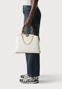 Bolso tote blanco con un patrón, asas de cuero marrón y un detalle de cadena, sostenido por una persona con jeans oscuros y zapatillas. Diseño texturizado visible.