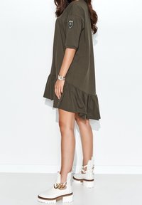 Vestido verde oliva de manga corta con dobladillo con volantes. Presenta una insignia en la manga. Combinado con botas blancas hasta el tobillo y un acento de cadena dorada.