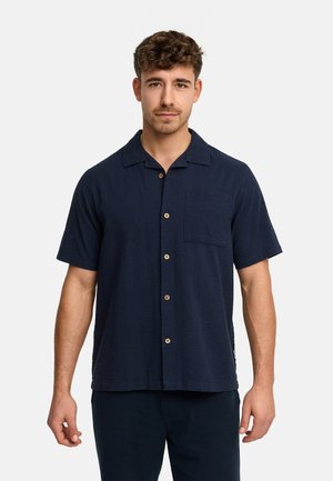 Mężczyzna ubrany w granatową, krótkorękawową koszulę na guziki z kieszenią na piersi, stoi na tle jednolitego, jasnoszarego tła.