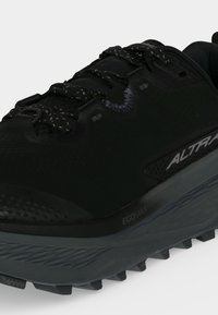 Gros plan d'une chaussure de trail noire avec des lacets à motifs, une semelle épaisse grise, et le logo "Altra" et "Ecomax" sur le côté.