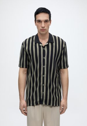 Muž s krátkymi tmavými vlasmi v čierno-olivovom vertikálne pruhovanom krátkym rukávom s gombíkmi a béžových nohaviciach na jednofarebnom pozadí.