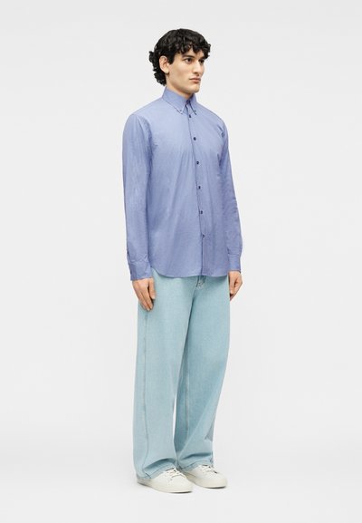 Νεαρός άνδρας που στέκεται φορώντας μπλε ριγέ πουκάμισο με κουμπιά, ανοιχτόχρωμα τζιν με φαρδιά μπατζάκια και λευκά αθλητικά παπούτσια σε απλό φόντο.