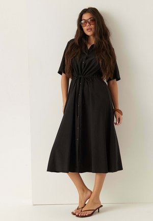 Femme aux longs cheveux portant de grandes lunettes de soleil, une robe midi noire boutonnée avec une taille à cordon, et des sandales marron à talons, debout contre un mur blanc.