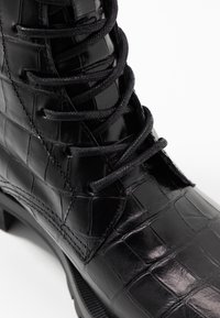 Botín de cuero negro con patrón de piel de cocodrilo, puntera redonda y cordones texturizados negros, sobre una suela de goma resistente.