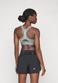 Sujetador deportivo verde con detalle recortado y tirantes ajustables, combinado con pantalones cortos de correr negros con costuras reflectantes y un logo redondo.
