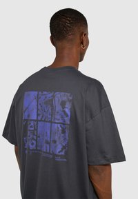 Czarny t-shirt z bawełny w kolorze ciemnoszarym z dużym, niebieskim nadrukiem graficznym na plecach, o kwadratowym wzorze i elementach tekstowych. Krótkie rękawy, luźny krój.