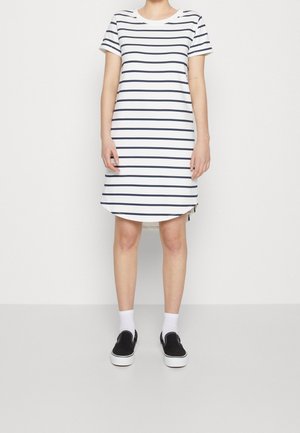Frau trägt ein lässiges weißes Kleid mit schwarzen horizontalen Streifen, kurzen Ärmeln, kombiniert mit weißen Socken und schwarzen Slippern.