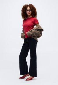 Γυναίκα με σγουρά μαλλιά που φορά γυαλιά, κόκκινο t-shirt, σκούρο τζιν με φαρδιά σκέλη, κόκκινες ψηλές γόβες και κρατά ένα καφέ μπουφάν περασμένο πάνω από τα χέρια της.