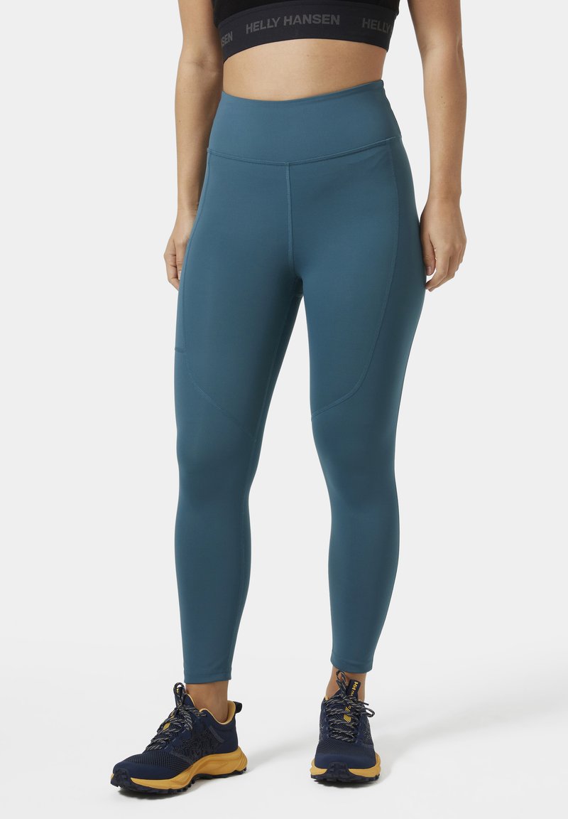 Teal sportlegging met hoge taille, gladde stof en zijzakken. Gecombineerd met donkerblauwe, gestructureerde hardloopschoenen.
