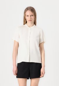 Short-sleeved, collared blouse in off-white fabric, featuring a button front and a straight cut. Paired with black shorts.