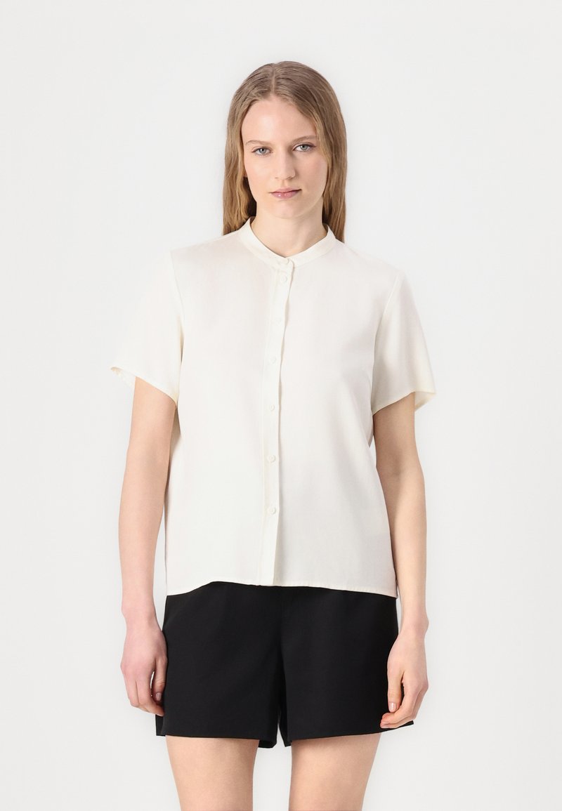 Short-sleeved, collared blouse in off-white fabric, featuring a button front and a straight cut. Paired with black shorts.