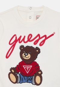 Camiseta branca de algodão com o logótipo "guess" bordado em vermelho, apresentando um urso castanho vestido com um suéter vermelho e umas calças de ganga azuis. Fechos de pressão no colarinho.