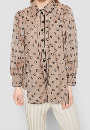 Blouse beige à manches longues avec motifs floraux noirs, boutons noirs et passepoil noir, portée avec un pantalon beige clair à rayures.