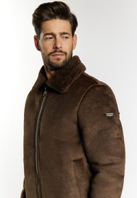 Barna shearling dzseki puha textúrával, cipzáros elülső résszel és dús gallérral. Oldalsó zsebekkel és testre szabott kialakítással rendelkezik. Látható márkajelzés nélkül.