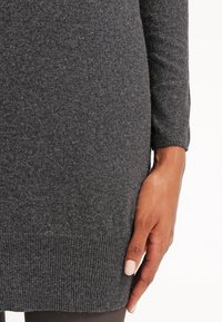 Gros plan sur la main d'une personne reposant sur un pull tricoté gris foncé à ourlet côtelé et manches longues.