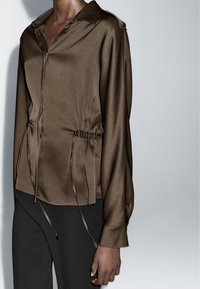 Bruine satijnen blouse met kraag, knoopsluiting aan de voorkant, aantrekkoord in de taille en lange mouwen, gecombineerd met zwarte broek. Gladde textuur en glans.