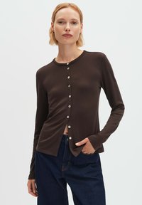 Frau mit kurzen blonden Haaren, trägt ein eng anliegendes, dunkelbraunes Langarm-Button-up-Oberteil und lockere dunkelblaue Jeans, eine Hand in der Tasche, neutraler Hintergrund.