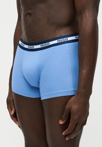 Torse et cuisses supérieures d'un homme portant un boxer bleu clair avec une ceinture bleu marine et blanche de marque "HUGO."