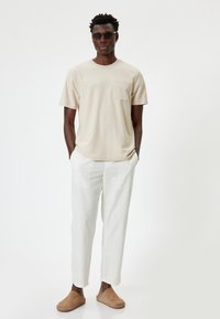 Beige bomulds T-shirt med brystlomme, bæret med hvide linnedbukser og brune ruskinds slip-ons. Modellen står med hænderne i lommerne.