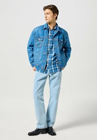Lichtblauwe denimjas met knoopsluitingen en borstzakken, gedragen over een blauw geruite shirt. Lichtblauwe jeans en zwarte laarzen.