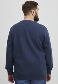 Felpa blu navy con scollo rotondo, polsini e orlo a costine. La vista posteriore mostra una texture del tessuto liscia e un design semplice e privo di decorazioni.