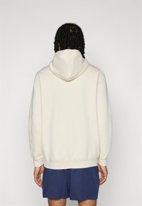 Haut à capuche crème avec une grande capuche, texture douce, poignets côtelés et coupe décontractée, associé à un short bleu marine.