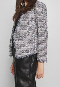 Donna che indossa un giacca di tweed blu e bianca con bordi frangianti e pantaloni di pelle neri a vita alta, inquadrata dalla nuca ai fianchi.