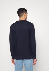 Marineblauwe textuurtrui met een ronde hals, lange mouwen en geribbelde naden; gedragen over lichtblauwe jeans.
