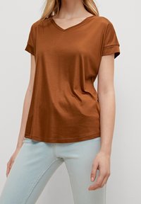 Braunes V-Ausschnitt-T-Shirt mit hochgekrempelten Ärmeln, aus einem glatten Stoff gefertigt, kombiniert mit hellblauen Jeans. Der Schnitt ist entspannt und leicht locker.