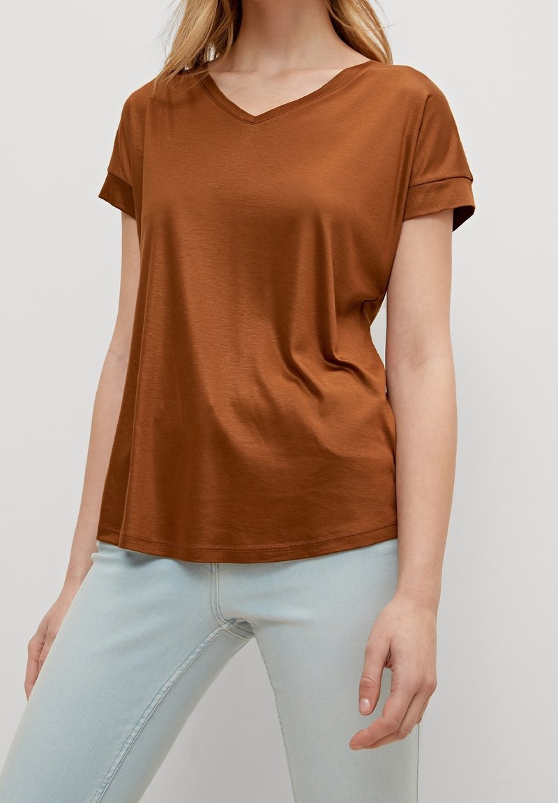 Braunes V-Ausschnitt-T-Shirt mit hochgekrempelten Ärmeln, aus einem glatten Stoff gefertigt, kombiniert mit hellblauen Jeans. Der Schnitt ist entspannt und leicht locker.