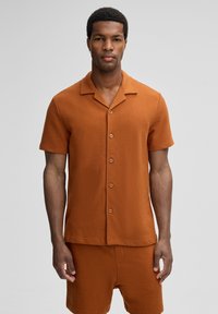 Uomo vestito con una camicia a maniche corte color ruggine e pantaloni corti abbinati, in piedi contro uno sfondo grigio uniforme.
