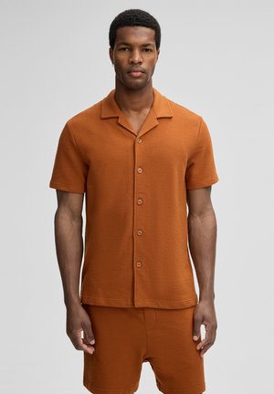 Man wearing a rust-colored short-sleeve button-up shirt and matching shorts, standing against a plain gray background.