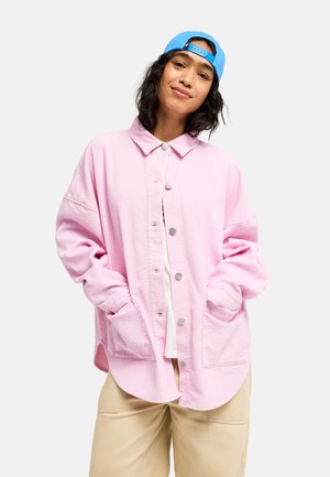Jeune femme aux cheveux foncés portant une casquette bleue à l'envers, une veste rose oversize boutonnée et un pantalon beige, souriante avec les mains dans les poches.