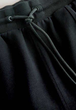 Mustad dressipüksid on valmistatud pehmest ja tekstuuriga kangast, millel on nööriga vöökoht ja kontrastsed metalldetailid kindlaks reguleerimiseks.