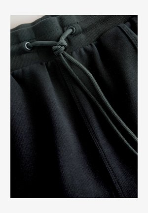 Mustad dressipüksid on valmistatud pehmest ja tekstuuriga kangast, millel on nööriga vöökoht ja kontrastsed metalldetailid kindlaks reguleerimiseks.